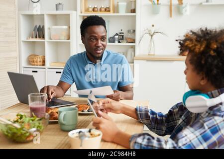 Afrikanischer junger Vater sitzt am Tisch vor dem Laptop und spricht mit seinem Sohn, während er in Tablet-pc spielt Stockfoto