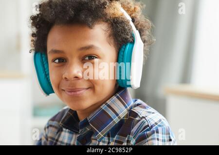 Porträt eines afrikanischen kleinen Jungen in Kopfhörern, der bei der Kamera lächelt, während er zu Hause sitzt Stockfoto