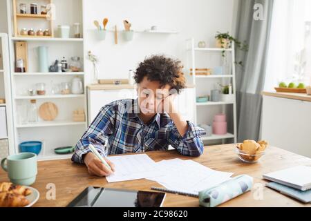 Afrikanischer Junge schreibt in seinem Notebook macht er seine Hausaufgaben, während er zu Hause am Tisch in der Küche sitzt Stockfoto