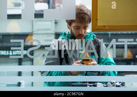 Deliveroo Take Away Mahlzeit Kurier Lieferung Radfahrer vor einem Restaurant, von wo er eine Sammlung macht. Er überprüft sein mobiles Gerät. England GB (120) Stockfoto