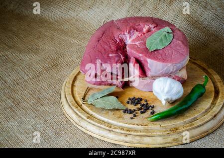 Großes, ungekohltes Stück rundes Rind auf Sackleinen mit Knoblauch und Gewürzen. Rohes Fleisch mit scharfer Paprika, Knoblauch, schwarzem Pfeffer und Lorbeerblättern auf einem Schneidebrett Stockfoto