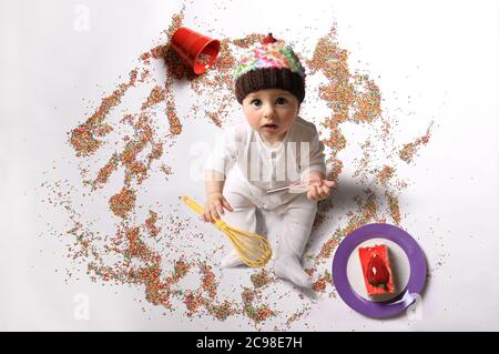 Ein Baby hält zwei Whisks und trägt einen Cupcake Hut zwischen Streuseln und ein Stück Kuchen. Stockfoto