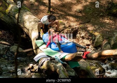 Paar Wanderer entspannen auf gefallenen Baumstamm über Berg Fluss Bach im sonnigen Sommer als shinrin Yoku Wald Baden und Natur Eskapismus. Stockfoto