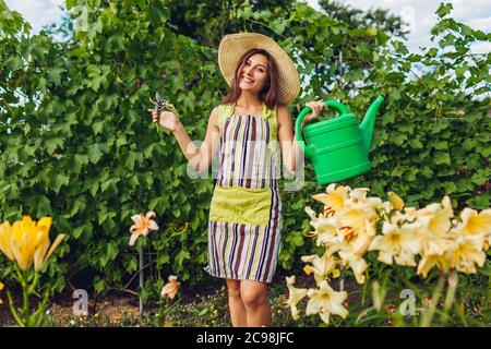 Frau, die Blumen mit Gießkanne gießt und im Garten mit dem Beschneider schneidet. Gärtner kümmert sich um Lilien. Stockfoto