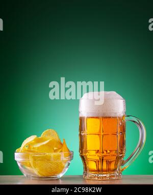 Becher helles Bier mit Schaum und Chips auf einem Teller auf grünem Hintergrund Stockfoto