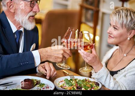 Schöne ältere Pärchen haben eine Mahlzeit in sympathisches Restaurant, verliebt. Grauhaarige männliche und weibliche Getränke- und Klappgläser mit Champagner. Stockfoto