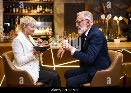 Reifes Paar kaukasischer Mann und Frau sitzen im Restaurant und essen Dessert, trinken ein Glas Wein. Männchen im Smoking, Weibchen im weißen Blazer. Frau hält Stockfoto