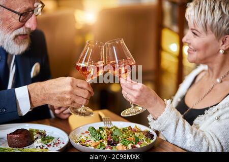 Schöne ältere Pärchen haben eine Mahlzeit in sympathisches Restaurant, verliebt. Grauhaarige männliche und weibliche Getränke- und Klappgläser mit Champagner. Stockfoto