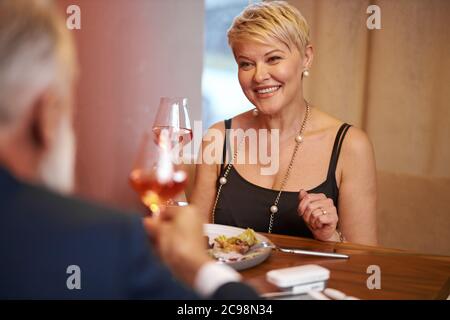 Kaukasische ältere Frau in schwarzem Kleid feines Abendessen Essen mit Mann in elegantem Restaurant. Weibchen hob ein Glas und ging zu trinken. Rückansicht auf Ehemann Stockfoto
