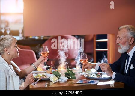 Ältere kaukasische Paar in der Liebe miteinander. Schöner Mann im Anzug und Frau in weißen clink Gläser mit Champagner. Leichter Rauch im Restaurant. Ros Stockfoto
