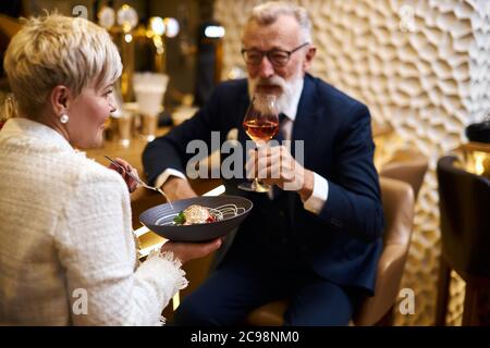 Reifes Paar kaukasischer Mann und Frau sitzen im Restaurant und essen Dessert, trinken ein Glas Wein. Männchen im Smoking, Weibchen im weißen Blazer. Frau hält Stockfoto