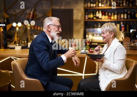 Reifes Paar kaukasischer Mann und Frau sitzen im Restaurant und essen Dessert, trinken ein Glas Wein. Männchen im Smoking, Weibchen im weißen Blazer. Frau hält Stockfoto