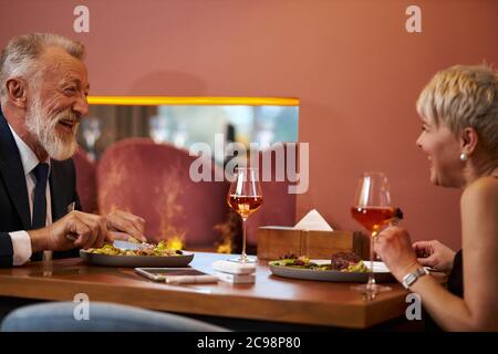 Ältere schöne Paar genießen Abend im Restaurant, schauen sich gegenseitig und lachen. Grauhaariger Mann im Smoking, attraktive Frau lächelt und lacht. Romant Stockfoto