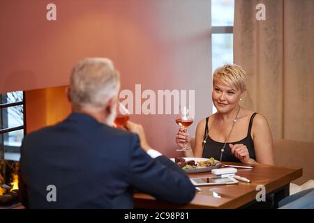 Kaukasische ältere Frau in schwarzem Kleid feines Abendessen Essen mit Mann in elegantem Restaurant. Weibchen hob ein Glas und ging zu trinken. Rückansicht auf Ehemann Stockfoto