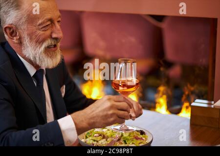 Eleganter älterer bärtiger und grauhaariger Mann im Smoking mit einem Glas Champagner sitzen im Restaurant und lachen, schauen Person gegenüber an. Hintergrund Kamin Stockfoto