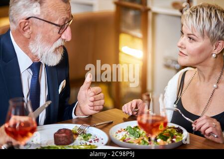 Hübsche Dame und älterer grauhaariger Mann im Smoking, der sich im Restaurant freundlich unternahm. Kollegen nach der Arbeit diskutieren und essen Stockfoto