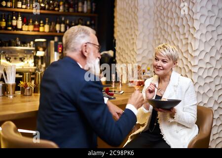 Reifes Paar kaukasischer Mann und Frau sitzen im Restaurant und essen Dessert, trinken ein Glas Wein. Männchen im Smoking, Weibchen im weißen Blazer. Frau hält Stockfoto