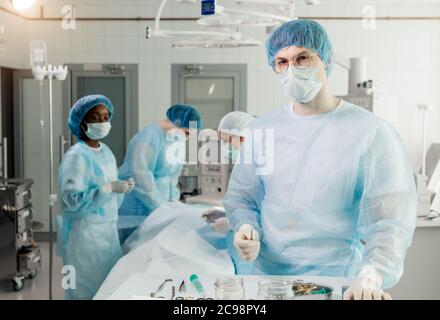 Professionelle qualifizierte Arzt mit Pinzette posiert, um die Kamera. Nahaufnahme Foto. Team im Hintergrund des photo.Handsome Chirurg mit einer Pause nach hinten Stockfoto