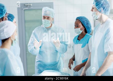 Cleverer Chirurg in Maske und Mütze, der seinen Mitarbeitern, die ihm sehr aufmerksam zulauschen, etwas erklärt. Stockfoto