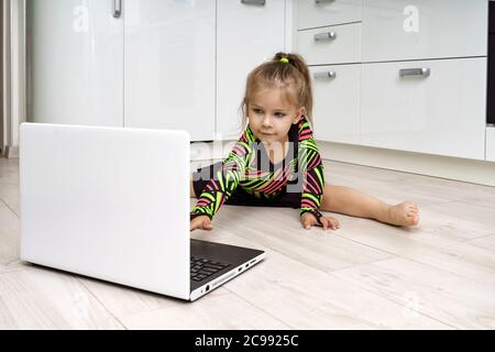 Kleines Mädchen in einem Turnanzug ist in rhythmische Gymnastik online zu Hause beschäftigt und schaut auf einen Laptop Stockfoto