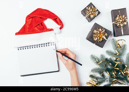 Frauenhandschrift in leerem Notizblock mit rotem Weihnachtsmann-Hut neben schwarzen Geschenkschachteln und verzierten Tannenzweigen auf weißem Tisch, flaches Lay. Stockfoto