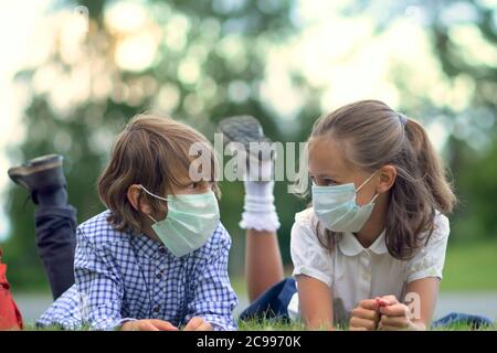 Die Kinder gehen zurück zur Schule. Nette Schüler mit Rucksäcken. Junge und Mädchen in Sicherheitsmasken liegen auf dem Gras. Stockfoto