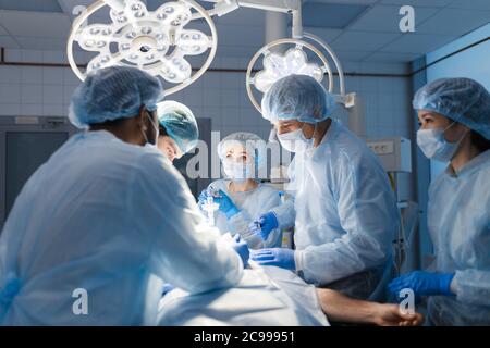 Multirassisches Team von Chirurgen, das sich während einer Herzoperation in einem Krankenhaus auf einen Patienten konzentriert. Reifer kaukasischer Arzt teilt seine Erfahrungen mit MUL Stockfoto