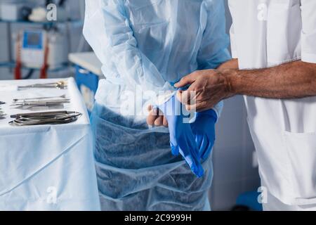 Unkenntlich chirurgischer männlicher Assistent hilft Chirurgen, Latexhandschuhe anzuziehen, während sie in der Chirurgie nah stehen. Stockfoto