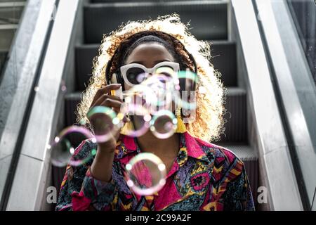 Anonyme afroamerikanische Frau in Kopfhörern und bunte Bluse weht transparente Blasen, während sitzen auf beweglichen Treppe im Rücken beleuchtet Stockfoto