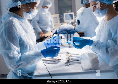 Zwei verschiedene weibliche Krankenschwestern, die Operationsinstrument für Gruppe von Chirurgen im Hintergrund operierenden Patienten in Operationssaal. Stockfoto