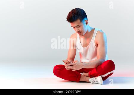 Junge sportliche stilvolle männliche Tänzerin in roten Jogginghosen Überprüfung seines Smartphones im Studio über weißem Hintergrund. Asiatische Hipster bboy lächelnd bei Kamera befo Stockfoto