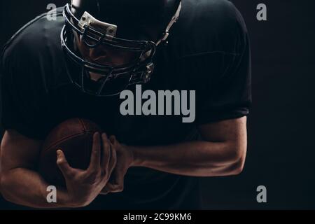 Schöner kaukasischer Mann in American Football Uniform und Helm läuft in Aktion mit einem Ball über dunklen Hintergrund, Nahaufnahme. Stockfoto