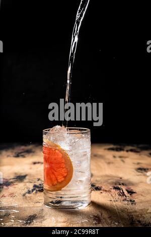 Gin und Tonic Cocktail Rezept, Gießen Tonic in ein Highballglas mit Eis gefüllt und garniert mit einer Grapefruit-Scheibe, Holztisch, dunkle Backgroun Stockfoto