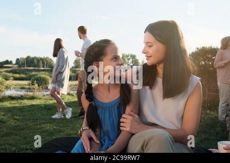 Junge Mutter umarmt und spricht mit ihrer kleinen Tochter, während sie draußen auf dem Land sitzt Stockfoto