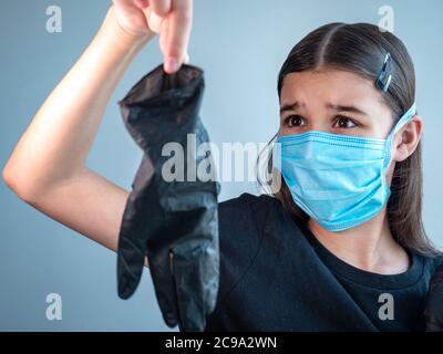 Verängstigte weibliche Teenager-Mädchen in einer blauen schützenden medizinischen Gesichtsmaske hält einen schwarzen Latex-Handschuh durch Spitzen ihrer Finger auf Armlänge. Coronavirus Stockfoto