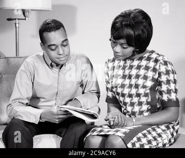 1960S AFRICAN-AMERICAN COUPLE SITTING ON COUCH EHEMANN SHOWING EHEFRAU ARTICLE IN NEWSPAPER SHE IS TRAGING DRESS HOUNDSTOOTH PRINT - N2234 HAR001 HARS LIFESTYLE FEMALS MARRIED STUDIO SHOT SHOWING EHEPARTNER HUSBANDS HEALTHINESS HOME LIFE COPY SPACE HALF-LENGTH LADIES PERSONS INSPIRATION FÜRSORGLICHE MÄNNER B&W PARTNER ERFOLG TRÄUME GLÜCK AFRI STILVOLLE PERSÖNLICHE BINDUNG ZUNEIGUNG EMOTION HAHNENTRITTKOPF MID-ADULT MANN MID-ADULT FRAU ZWEISAMKEIT FRAUEN SCHWARZ UND WEISS HAR001 ALTMODISCH Stockfoto