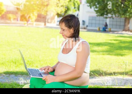 Glückliche schwangere Frau, die an einem sonnigen Tag im Freien auf einer grünen Wiese in einem Stadtpark sitzt Stockfoto