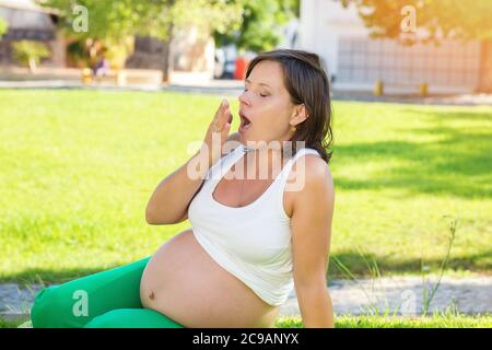 Schwangere Frau gähnend. Schöne Zukunft Mutter bereitet sich auf Geburt des Babys, müde und versuchen, sich zu entspannen, sitzen auf einem grünen Gras in einem sonnigen Park im Freien Stockfoto