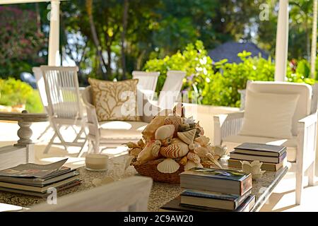 Couchtisch mit Büchern und Muscheldekor, mit Stühlen und Esstisch, auf einer schattigen Veranda mit einem sonnendurchfluteten Garten dahinter auf Barbados Stockfoto