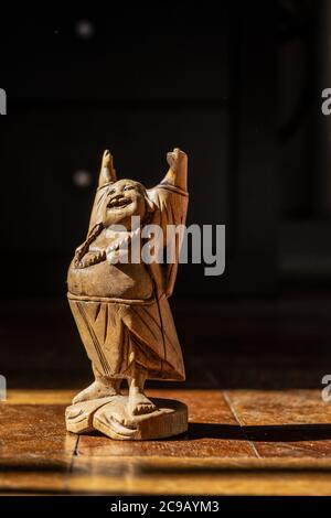 Holzstatue eines glücklichen buddhistischen Mönchs, der in einem Sonnenfleck auf einem abgenutzten Holzparkettboden steht. Stockfoto