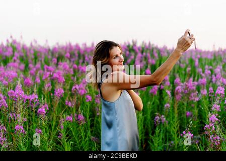 Frau in grauem Kleid macht Selfie Foto auf Handy auf Feuerweed Wiese Stockfoto