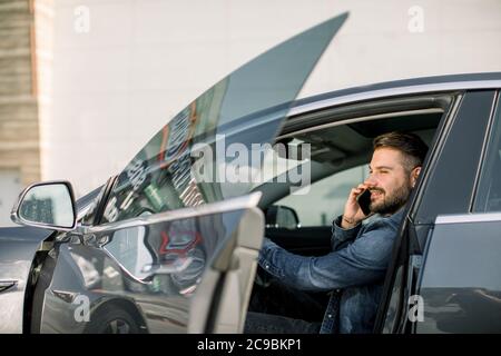 Bärtiger junger Geschäftsmann in legerer Jeans-Jacke, mit Handy, in seinem modernen Luxuswagen vor dem Bürogebäude der Stadt oder beim Einkaufen Stockfoto