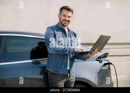 Junger, gutaussehender lächelnder Mann, der am Laptop arbeitet, wartet, während sein Luxus-Elektroauto charchiert, für das Aufladen bezahlt und in der Nähe des Bahnhofs steht Stockfoto