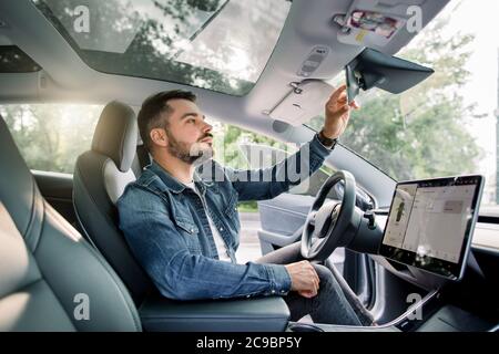 Junge kaukasische Mann trägt lässige Jeans-Shirt, sitzt in seinem neuen elektrischen autonomen Auto-Lenkung, Blick in den Auto-Spiegel innen, warten Stockfoto