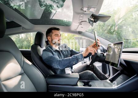 Hübscher kaukasischer Mann, der in einem modernen selbstlenkenden Elektrofahrzeug sitzt, mit einer mobilen App auf seinem Smartphone, während er geparkt ist, lädt am Kompressor auf Stockfoto
