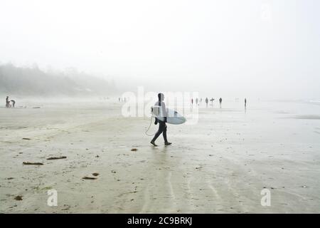 Surfer im Neoprenanzug mit einem Surfbrett am Cox Bay Beach in Tofino, British Columbia, Kanada. Nebliger Tag am Strand in Tofino, BC, Kanada Westküste. Stockfoto