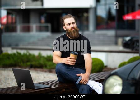 Crop junge bärtige Mann studieren oder arbeiten mit Laptop halten Take-away Tasse Kaffee, während auf der Bank sitzen und auf die Kamera Stockfoto