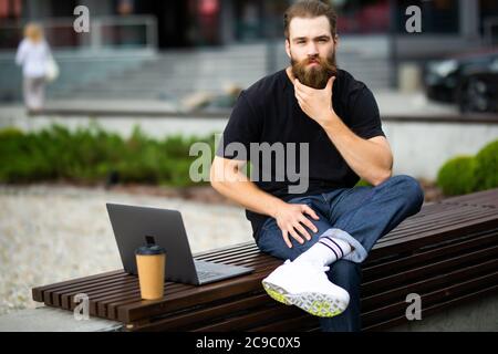 Porträt eines jungen Mannes mit Laptop und eine Tasse Kaffee auf der Bank im Park Stockfoto