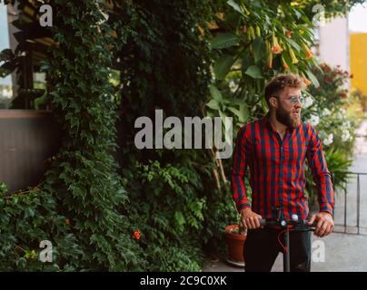 Hübscher junger bärtiger Kaukasusmann mit Sonnenbrille reitet auf dem Street e Scooter Stockfoto