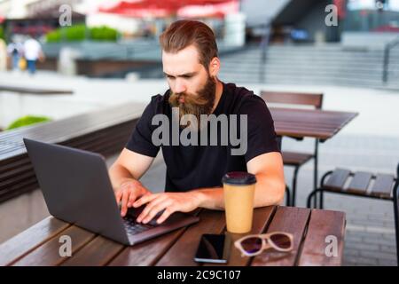 Junger Mann, der eine Brille trägt und eine Tasse Kaffee hält und auf dem Laptop tippt, während er im Café im Freien arbeitet Stockfoto